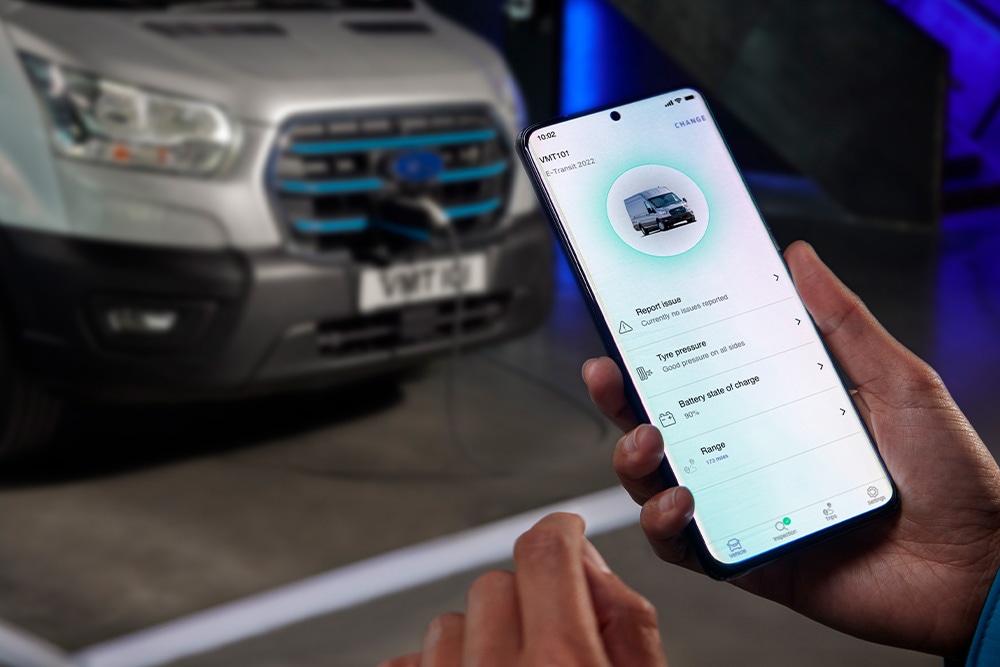 Een persoon houdt een smartphone vast waarop een app-interface wordt weergegeven, met een zilveren Ford Pro Transit bestelwagen wazig op de achtergrond.