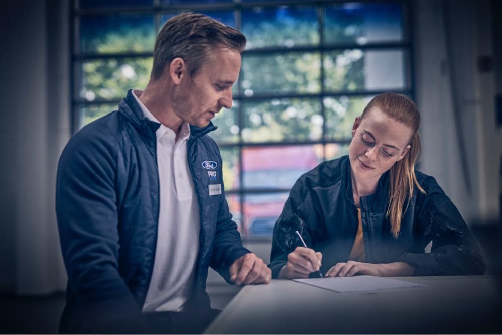 Twee Ford Pro medewerkers aan een tafel, één ondertekent een document, de ander observeert in een werkplaats.