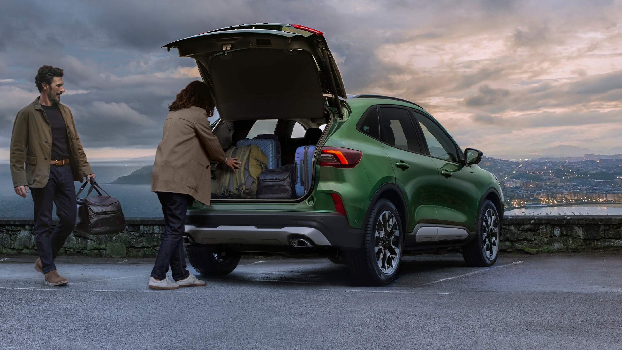 Een man en een vrouw laden bagage in de kofferbak van een groene SUV die geparkeerd staat op een verharde uitkijkplaats.