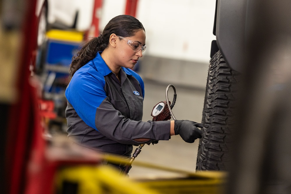 Een Ford-technicus met veiligheidsbril en handschoenen controleert de bandenspanning van een voertuig. 